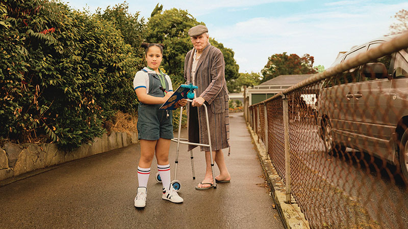 A young girl with a clipboard and her elderly neighbour with a walker in a driveway, staring at the viewer.